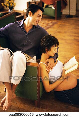 Stock Photograph - Man sitting backwards on chair and pregnant woman sitting on floor, reading and leaning head on man, portrait. Fotosearch