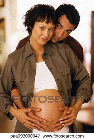Stock Photo - Man touching pregnant woman's stomach from behind, looking at camera, portrait. Fotosearch