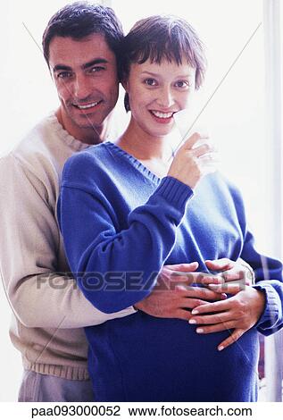 Stock Image - Man with arms around pregnant woman, portrait. Fotosearch