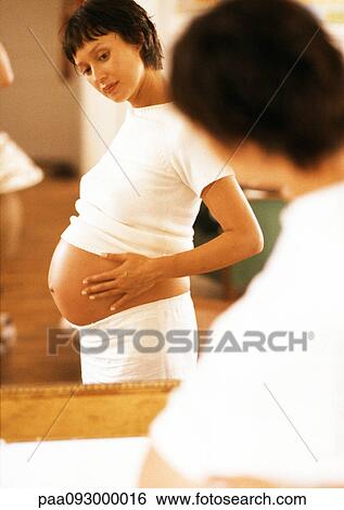 Stock Photograph - Pregnant woman looking at her stomach in mirror. Fotosearch