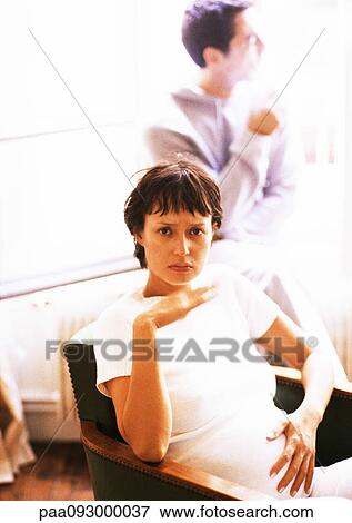 Stock Photo - Pregnant woman sitting in armchair, man in background. Fotosearch