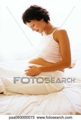 Pregnant woman sitting on bed, looking at her stomach View Large Photo Image Stock Image - Pregnant woman sitting on bed, looking at her stomach. Fotosearch