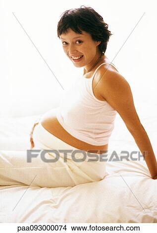 Pregnant woman sitting on bed, smiling at camera, portrait View Large Photo Image Picture - Pregnant woman sitting on bed, smiling at camera, portrait. Fotosearch