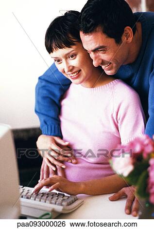 Stock Photograph - Pregnant woman using computer, man with arms around her looking over her shoulder. Fotosearch