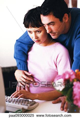 Stock Photography - Pregnant woman using computer, man with arms around her looking over her shoulder. Fotosearch