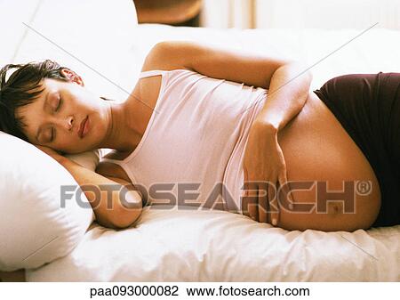 Stock Image - Pregnant woman with eyes closed, lying on side on bed. Fotosearch
