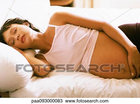 Stock Image - Pregnant woman with eyes closed, lying on side on bed. Fotosearch