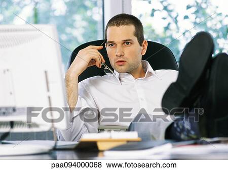 Businessman holding glasses next to face, looking at computer screen, sitting with feet on desk View Large Photo Image Stock Photo - Businessman holding glasses next to face, looking at computer screen, sitting with feet on desk . Fotosearch