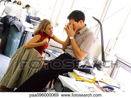 Stock Photo - In office, woman gesturing and talking to man, man sitting on desk with hands in front of face. Fotosearch