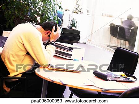 Stock Image - Man sitting at table, holding head with hands in office. Fotosearch