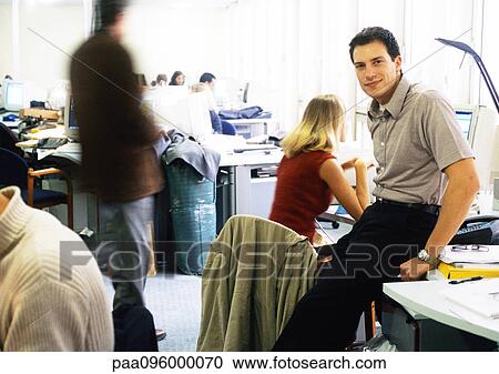 Stock Image - Man sitting on desk in office, looking at camera. Fotosearch