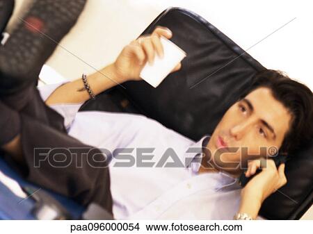 Picture - Man sitting with feet up, talking on telephone and holding plastic cup. Fotosearch