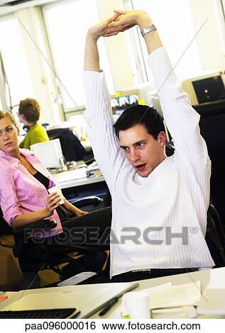 Stock Photograph - Man stretching in office, colleagues in background. Fotosearch