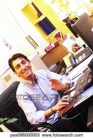 Stock Image - Man working at desk in office, smiling at camera. Fotosearch
