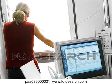 Stock Image - Woman in office standing and looking out of window, computer monitor in foreground. Fotosearch