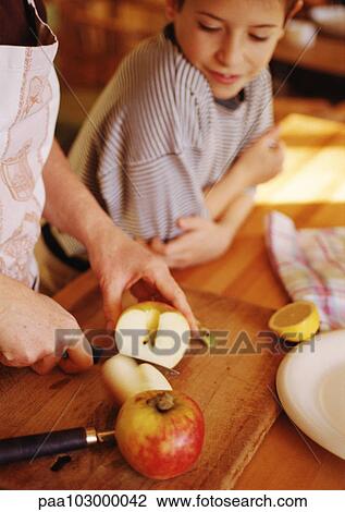 Stock Image - Hands cutting apples, child leaning on counter watching. Fotosearch