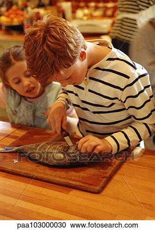 Stock Image - Two children in kitchen, one cutting a fish, parent in background. Fotosearch