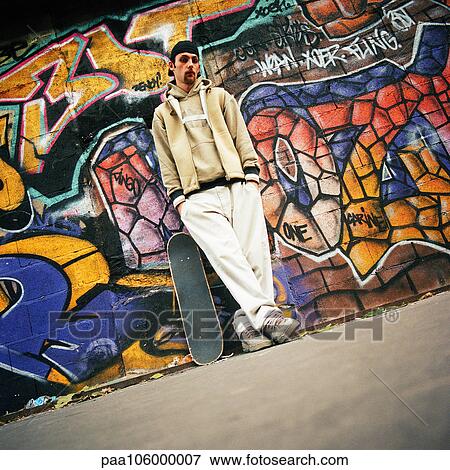 Stock Photo - Young man with skateboard leaning against graffitied wall, portrait. Fotosearch