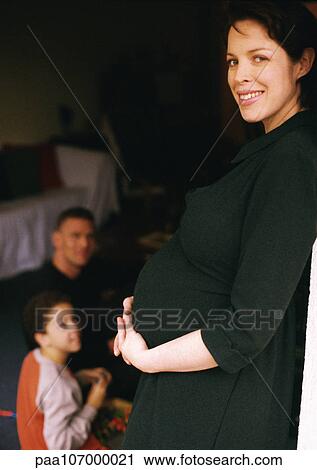 Stock Image - Pregnant woman standing and holding stomach, man and child sitting in background. Fotosearch