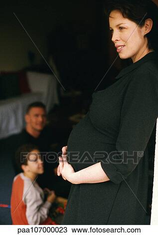 Pregnant woman standing and holding stomach, man and child sitting in background View Large Photo Image Stock Image - Pregnant woman standing and holding stomach, man and child sitting in background. Fotosearch