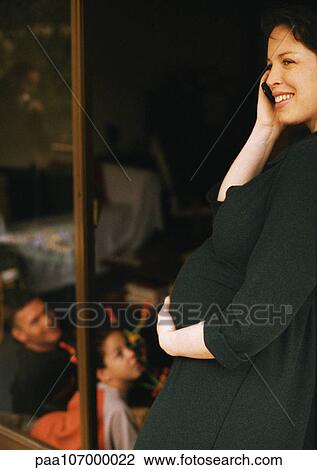 Stock Image - Pregnant woman standing, using cell phone and holding stomach, man and child sitting in background. Fotosearch