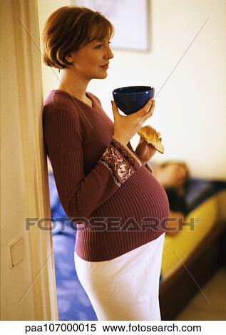 Stock Photography - Pregnant woman standing with bowl and croissant, side view. Fotosearch