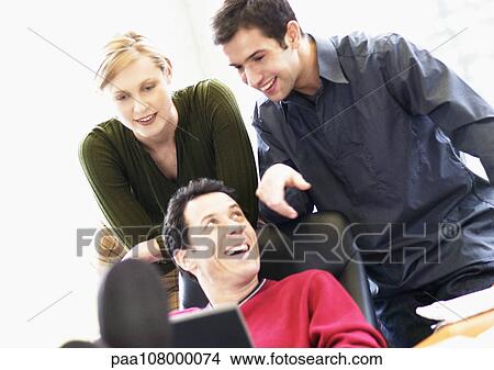 Picture - Man sitting in chair, second man and woman looking over his shoulder. Fotosearch