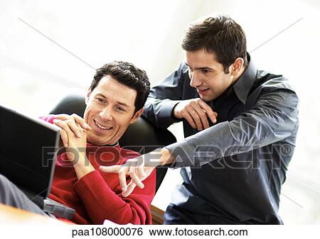 Stock Photograph - Man sitting in chair using laptop, second man looking over his shoulder, pointing. Fotosearch