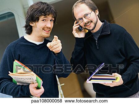 Two men looking into camera, one pointing, the other using cell phone, portrait View Large Photo Image Stock Photo - Two men looking into camera, one pointing, the other using cell phone, portrait. Fotosearch