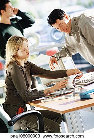 Stock Image - Woman sitting at desk looking at document, man leaning toward her, second man talking on phone in background. Fotosearch