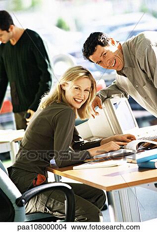 Woman sitting at desk, man leaning over desk, both smiling at camera View Large Photo Image Picture - Woman sitting at desk, man leaning over desk, both smiling at camera. Fotosearch