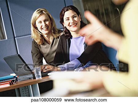 Women sitting at laptop computer, smiling, person pointing in foreground, blurred View Large Photo Image Stock Photo - Women sitting at laptop computer, smiling, person pointing in foreground, blurred. Fotosearch