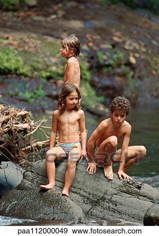 Stock Photo - Boy and two girls in swimming trunks on a big rock, outside. Fotosearch