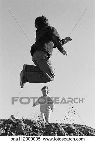 Stock Photography - Boy jumping and little girl walking in background, low angle view, b&w. Fotosearch