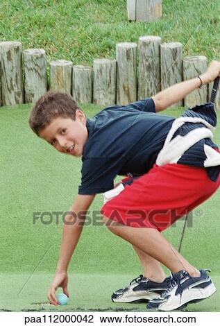 Stock Image - Boy standing on grass, picking up golf ball. Fotosearch