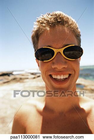 Stock Image - Close up of teenage boy wearing sunglasses at the beach.. Fotosearch