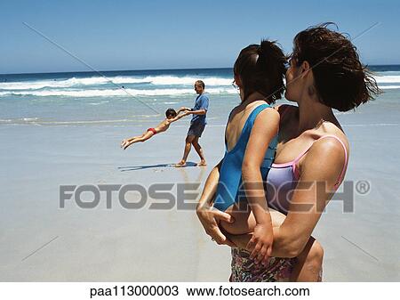 Stock Image - Woman holding little girl in arms in foreground on beach, man swinging little boy in background. Fotosearch