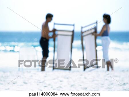 Picture - Two adults holding folded beachchairs on beach, blurred. Fotosearch