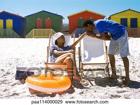Stock Photo - Woman sitting on beach, man kissing her hand. Fotosearch