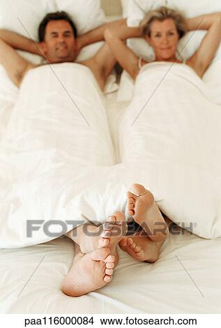 Picture - Couple lying in bed with arms behind heads, focus on feet in foreground. Fotosearch