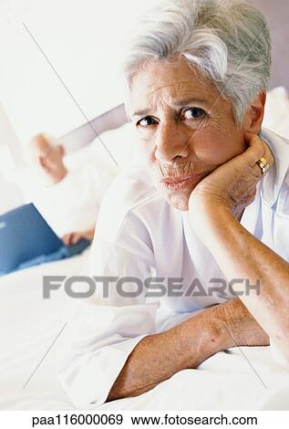 Mature woman on bed with hand under chin, man using laptop in background View Large Photo Image Stock Photo - Mature woman on bed with hand under chin, man using laptop in background. Fotosearch