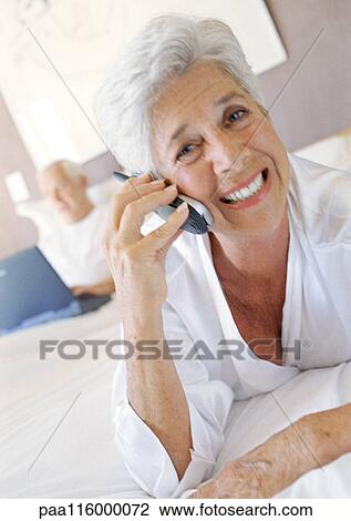 Mature woman using cell phone, man using laptop in bed in background View Large Photo Image Stock Image - Mature woman using cell phone, man using laptop in bed in background. Fotosearch