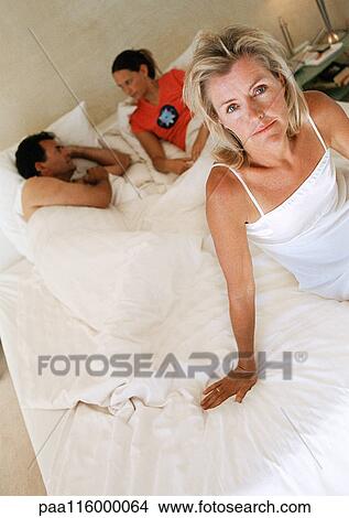 Picture - Woman sitting on bed, two people lying in bed in background. Fotosearch