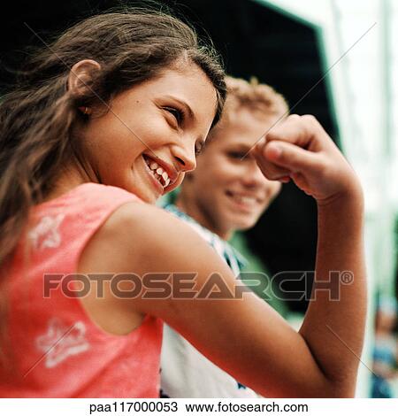 Stock Image - Girl flexing arm muscles, boy in background, smiling. Fotosearch