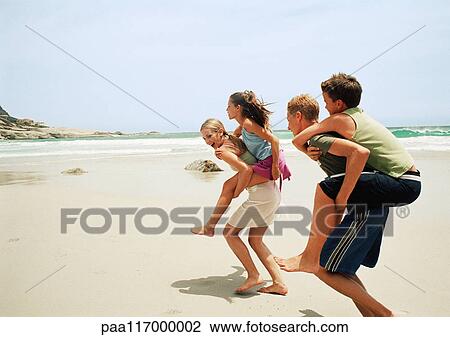 Stock Image - Two young people carrying children piggyback on the beach. Fotosearch