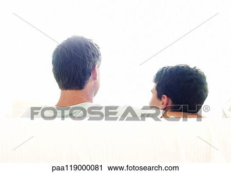 Stock Image - Man and son sitting on sofa, rear view. Fotosearch