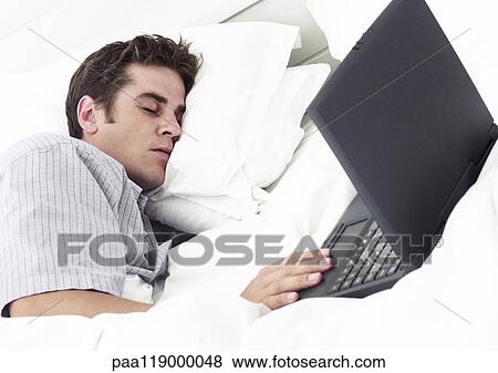 Stock Photo - Man lying on bed with eyes closed, hand on laptop computer. Fotosearch