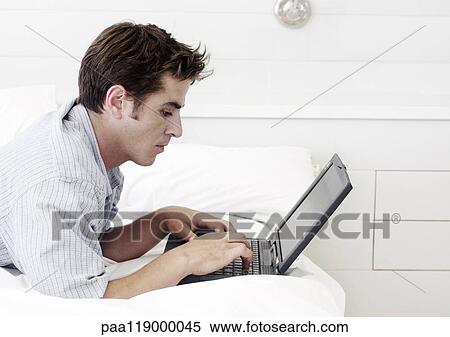 Stock Photography - Man lying on stomach, using laptop computer, side view. Fotosearch