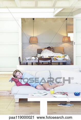 Stock Image - Woman lounging on sofa, man lying in bed in background. Fotosearch