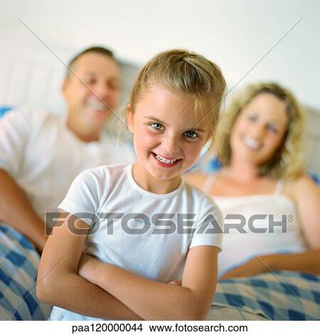 Girl folding arms and smiling in front of parents in bed, portrait View Large Photo Image Picture - Girl folding arms and smiling in front of parents in bed, portrait. Fotosearch